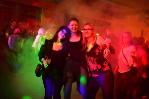 Drei lachende Frauen posieren bei einer Party, im Hintergrund Nebel und farbige Lichtakzente; Getränke und festliche Kleidung.