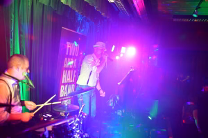 Musiker auf der Bühne singt und spielt Gitarre, hinter ihm farbiges Licht und Banner; Hintergrund zeigt Schlagzeug und Tonanlage.