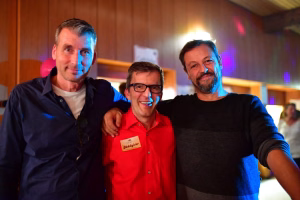 Drei Personen posieren lächelnd eng beieinander in warmem Licht, im Hintergrund Partydeko und farbige Bühnenbeleuchtung.