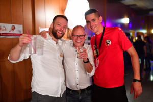 Drei Personen stoßen beim Fest an, eine Person hält ein Getränk; fröhliche Gesichter, Partylicht im Hintergrund, Zweck: gemeinsames Feiern.