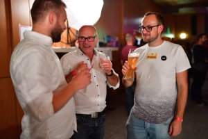 Drei Personen stoßen in geselligem Raum mit Bier an; Gläser sind erhoben, freundliche Stimmung bei einer Feier.