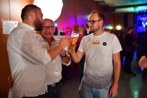 Drei Männer stoßen in einer festlich beleuchteten Halle mit Gläsern an und feiern gemeinsam.