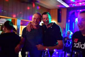 Zwei Männer posieren lachend nebeneinander bei einer Feier mit farbigem Bühnenlicht und ausgeblendeter Party-Umgebung im Hintergrund.