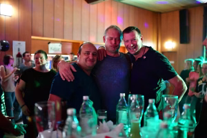 Drei Männer feiern eng beieinander lächelnd in einer festlichen Gesellschaft, ein Arm liegt freundschaftlich um die Schultern.
