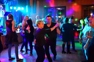 Menschen tanzen auf einer Partyfläche unter farbigem Licht, ein Paar in der Vordergrundposition hebt die Arme; Tanzfläche mit Publikum im Hintergrund.