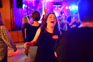 Person singt laut beim Feiern auf einer Bühne, im Vordergrund, mit buntem Licht und Publikumshintergrund.