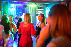 Menschen tanzen auf einer Party in beleuchtetem Saal; farbige Lichtstimmung und Bühne im Hintergrund schaffen festliche Atmosphäre.