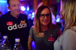 Drei Personen lachen im Event-Licht; im Vordergrund Getränke und verschwommene Hintergrunddeko, Szene in gemütlicher Feieratmosphäre.