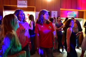 Party-Szene in beleuchtetem Innenraum: mehrere Gäste tanzen, bewegen sich im Licht von Blau und Rot; Schilder weisen auf Getränke hin.