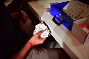 Hand hält Eintrittskarten in einer Abendveranstaltung; im Hintergrund Blauer Tisch, Karten und Spielmarker zur Besucherinformation.