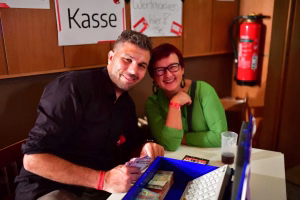 Zwei Personen lächeln am Tisch, mit blauem Display und Getränk; im Hintergrund Schilder und ein roter Feuerlöscher als Event-Umgebung.