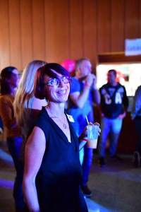 Teilnehmende Person in dunklem Kleid mit Brille, lächelnd und mit Getränk sowie Trinkhalm; im Hintergrund Veranstaltungsgäste und Projektion.