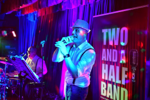 Sänger im weißen Hemd mit Mikrofon vor Bühne, umgeben von bunten Scheinwerfern und großem Schriftbanner „TWO AND A HALF“.
