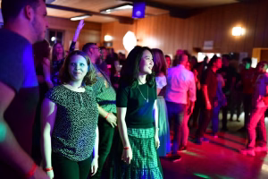 Zwei Frauen stehen im Vordergrund bei einer Feier, beleuchtet von Bühnenlicht; im Hintergrund tanzt und lächelt eine Gruppe im Saal.