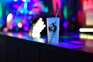 Auf einem Tresen steht ein Partybecher mit Trinkhalm; im Hintergrund buntes Bühnenlicht und unscharfe Dekoration zur Feier.