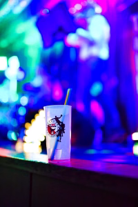 Becher auf einer Theke mit Trinkhalm; unscharfes Bühnenensemble im farbigen Partylicht, verdeutlicht das Fest-Atmosphäre.