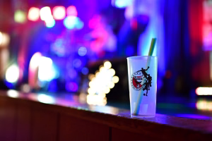 Plastikbecher auf einer Theke bei farbigem Bühnenlicht, unscharfer Hintergrund mit Lichtern zur Veranstaltungspromotion.