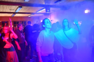 Feiernde Gäste auf einer Tanzfläche in buntem Licht, im Hintergrund eine Bar/Empore; die Szene zeigt ausgelassene Stimmung bei einer Veranstaltung.