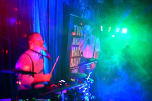 Live-Bühne mit zwei Musikerinnen im Spotlight; eine singt am Mikrofon, die andere spielt Gitarre, farbige Lichtnebel und Vorhang im Hintergrund.