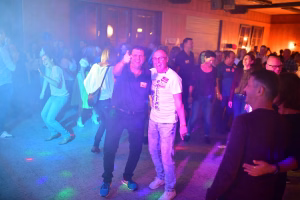 Zwei Männer lachen auf einer Tanzfläche bei Partylicht; im Hintergrund feiern Gäste in warmem Bühnenraum.