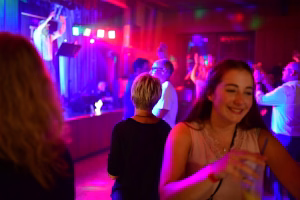Besucherinnen und Besucher bei einer Feier auf der Tanzfläche; im Hintergrund beleuchtete Bühne mit Musikern, farbiges Licht erzeugt Partystimmung.