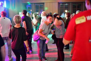 Menschen tanzen auf einer beleuchteten Feier im Innenraum; farbige Lichtfarben, Publikum im Hintergrund, freundliche Stimmung.
