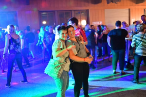 Zwei feiernde Frauen mit Einkaufsbeuteln umarmend, während im Hintergrund eine Partygesellschaft im Saal bei farbigem Licht tanzt
