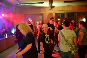 Feiernde Personen in einer Halle, beleuchtet von bunten Partylichtern; mehrere Gäste tanzen und stoßen mit Getränken an.