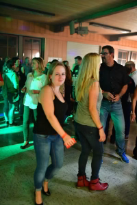 Menschen stehen auf einer beleuchteten Tanzfläche; zwei Frauen und ein Mann posieren im Vordergrund bei einer geselligen Feier in der Halle.