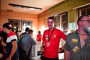 Mann im roten Vereinsshirt steht in einer Halle und hält eine Flasche; im Hintergrund Gespräche bei einer Veranstaltung.