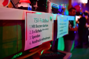 Hinweisschild mit Text „TSV-Fotobox“ und Schritten „Mit Buzzer starten“, „lächeln“, „Spenden“, „Ausdruck mitnehmen“ bei Festbeleuchtung.