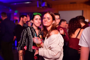Zwei Frauen bei einer Party im Partyraum, eine hält ein Glas, Konfetti-Atmosphäre mit farbigem Licht im Hintergrund.