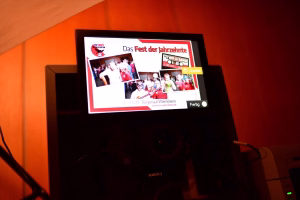 Digitales Display auf einer Bühne mit Folie „Das Fest der Jahrzehnte“ und Collage mehrerer Gruppenfotos, als Präsentation.