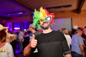 Mann mit farbenfroher Perücke und Partybrille im Innenraum, gibt Daumen hoch; wirkt als Teilnehmer bei einer Feier.