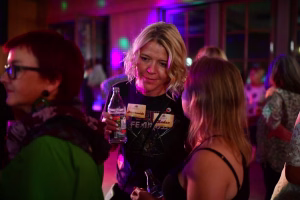 Frau in Partybeleuchtung hält ein Glas und lächelt, während andere Gäste im Hintergrund sitzen; Szene wirkt gesellig und feierlich.