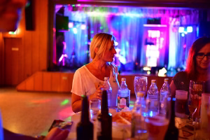 Frau steht neben einer gedeckten Bar mit Flaschen und Gläsern; im Hintergrund farbiges Bühnenlicht, Stimmung bei einer Feier.