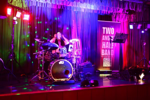 Musiker auf einer Bühne am Schlagzeug, grelles Farblicht in Lila/Grün, Vorhang und Beschriftung „Two and a Half Band“ im Hintergrund.