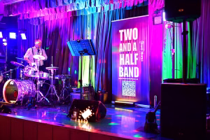 Bühne mit Gitarrist, Mikrofon und Schlagzeug; farbige Beleuchtung und leuchtendes Band-Logo-Display mit Text sowie QR-Code im Hintergrund.