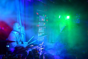 Musiker am Keyboard auf einer Bühne, farbiges Licht (blau/grün/rot) und Nebel, Live-Konzertstimmung im Hintergrund.