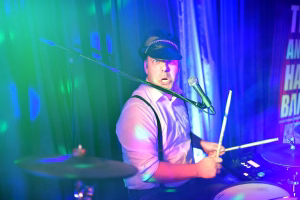 Musizierender Schlagzeuger am Mikrofon mit Kopfhörer und Kappe, spielt mit Sticks; Bühnenlicht in Blau und Grün als Bühnenevent.