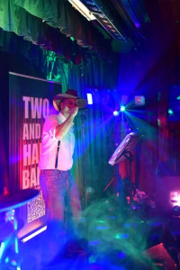 Sänger auf der Bühne mit Mikrofon, Hut und modischem Outfit; farbige Scheinwerfer und Vortragswand im Hintergrund bei Konzert-Atmosphäre.