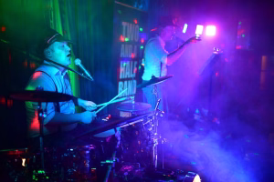 Musiker mit Hut am Mikrofon und Schlagzeuger im Hintergrund bei Live-Konzert, farbiges Bühnenlicht und Nebel.