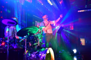 Sänger auf einer Bühne mit Mikrofon und Gitarre, beleuchtet von farbigem Bühnenlicht; im Vordergrund Drums und Mikroständer.