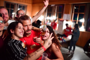 Gruppe feiert in einer Festhalle, macht Selfie, eine Person hebt den Arm, alle schauen zur Kamera; Gläser und Partyatmosphäre.