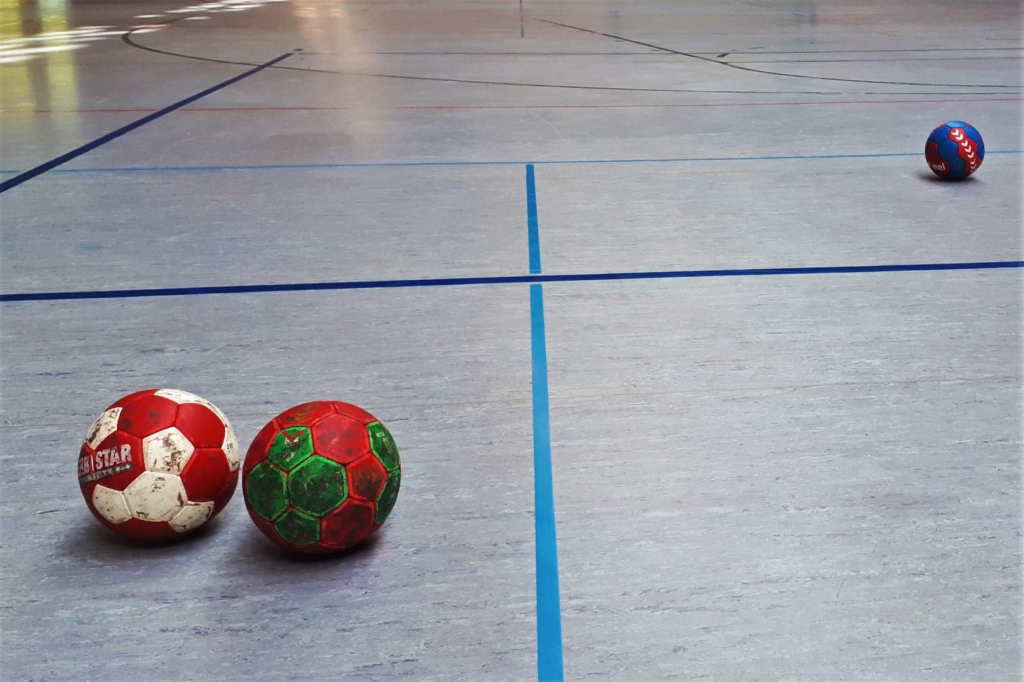 Drei Handbälle auf einem Hallenboden entlang von Linien: einer rechts, zwei vorne – verdeutlichen Spielfeld und Zielbereich.