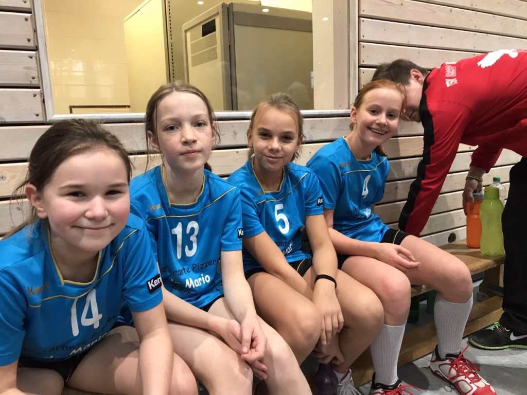 Vier Kinder in blauen Trikots sitzen eng auf einer Bank im Innenraum einer Sporthalle; daneben bereiten sich Betreuer vor.
