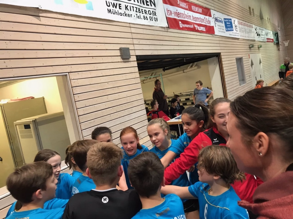 Innenansicht einer Sporthalle: Jugendliche um eine Bank versammelt, schauen nach vorn und wirken bei einem Spieltagstreffen konzentriert.