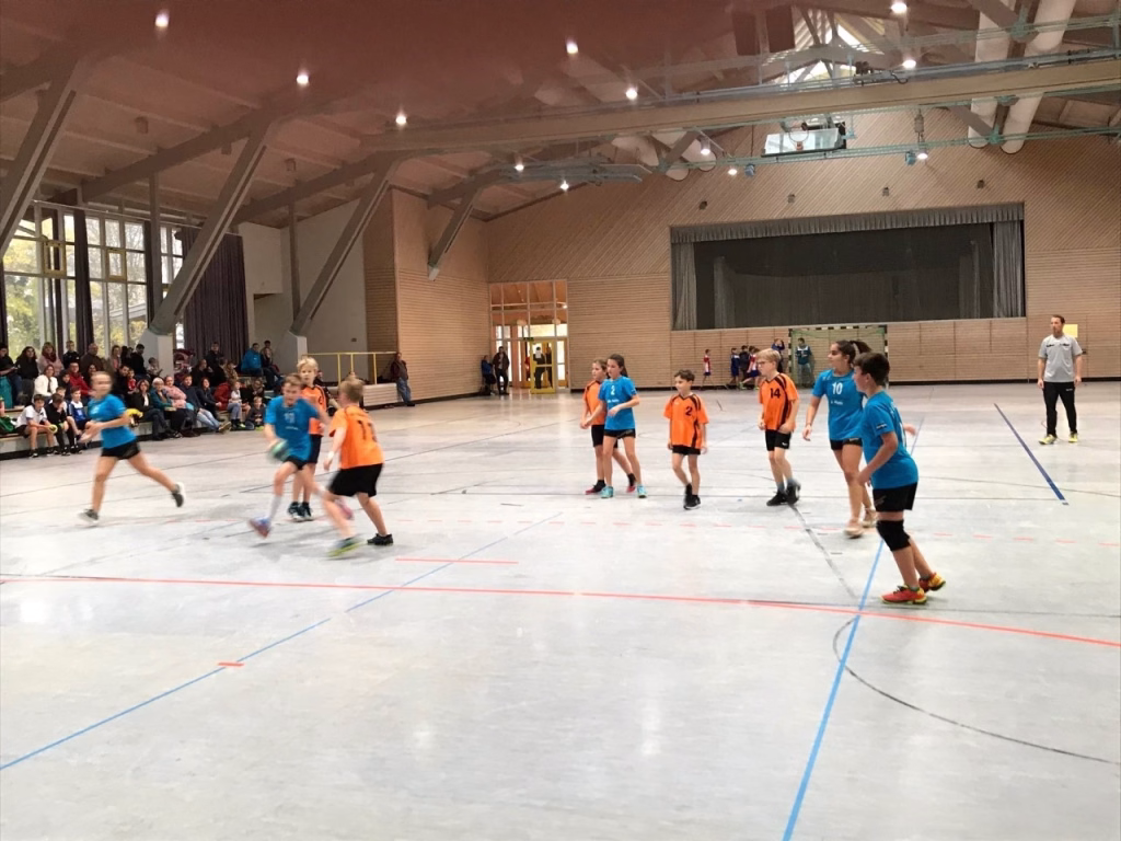 Handballspiel in der Sporthalle: Kinder in zwei Trikots laufen und werfen, Zuschauer sitzen an der Seitenlinie.