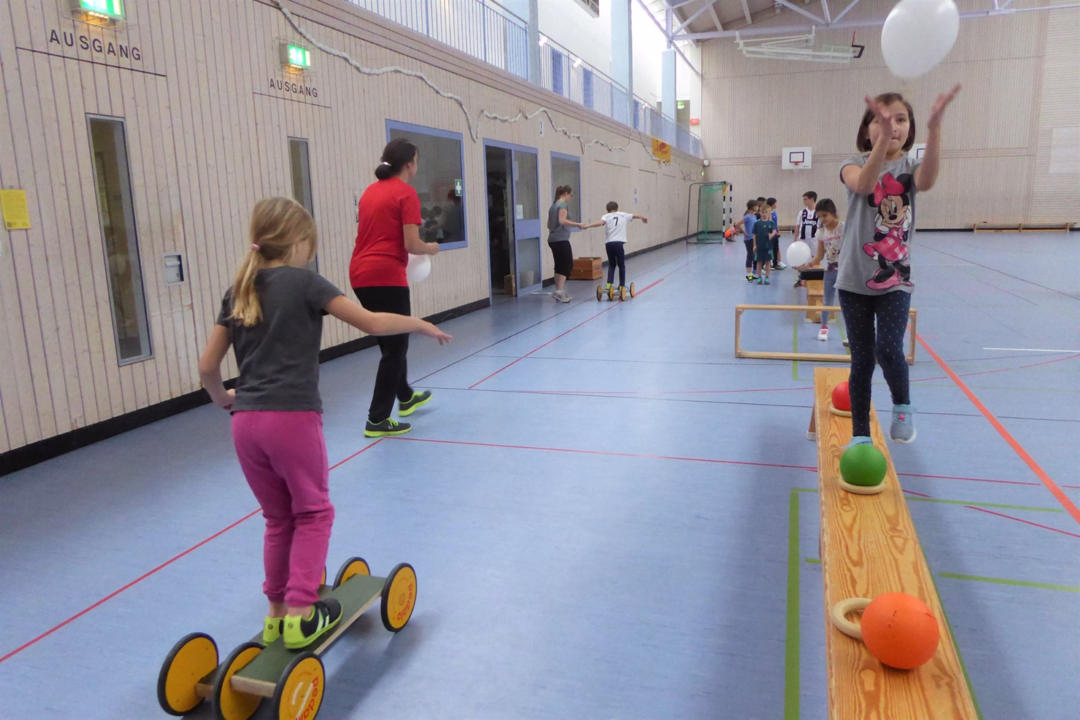 Turnhalle beim Aktionstag: Kinder balancieren auf Schwebebalken und stehen auf Skateboards mit Wellen; Sport und Geschicklichkeit.