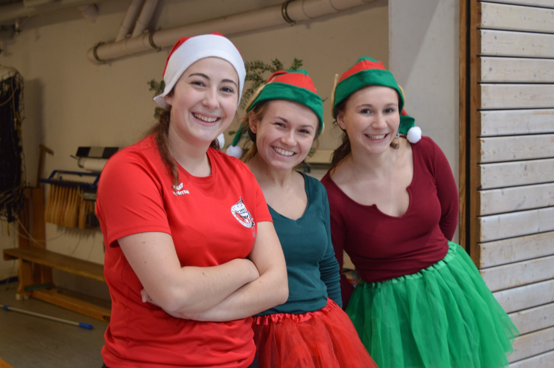 Drei Frauen lächeln in Weihnachtskostümen mit roten und grünen Mützen, aufgenommen in einer Turnhalle; fröhlicher Gemeinschaftsauftritt.